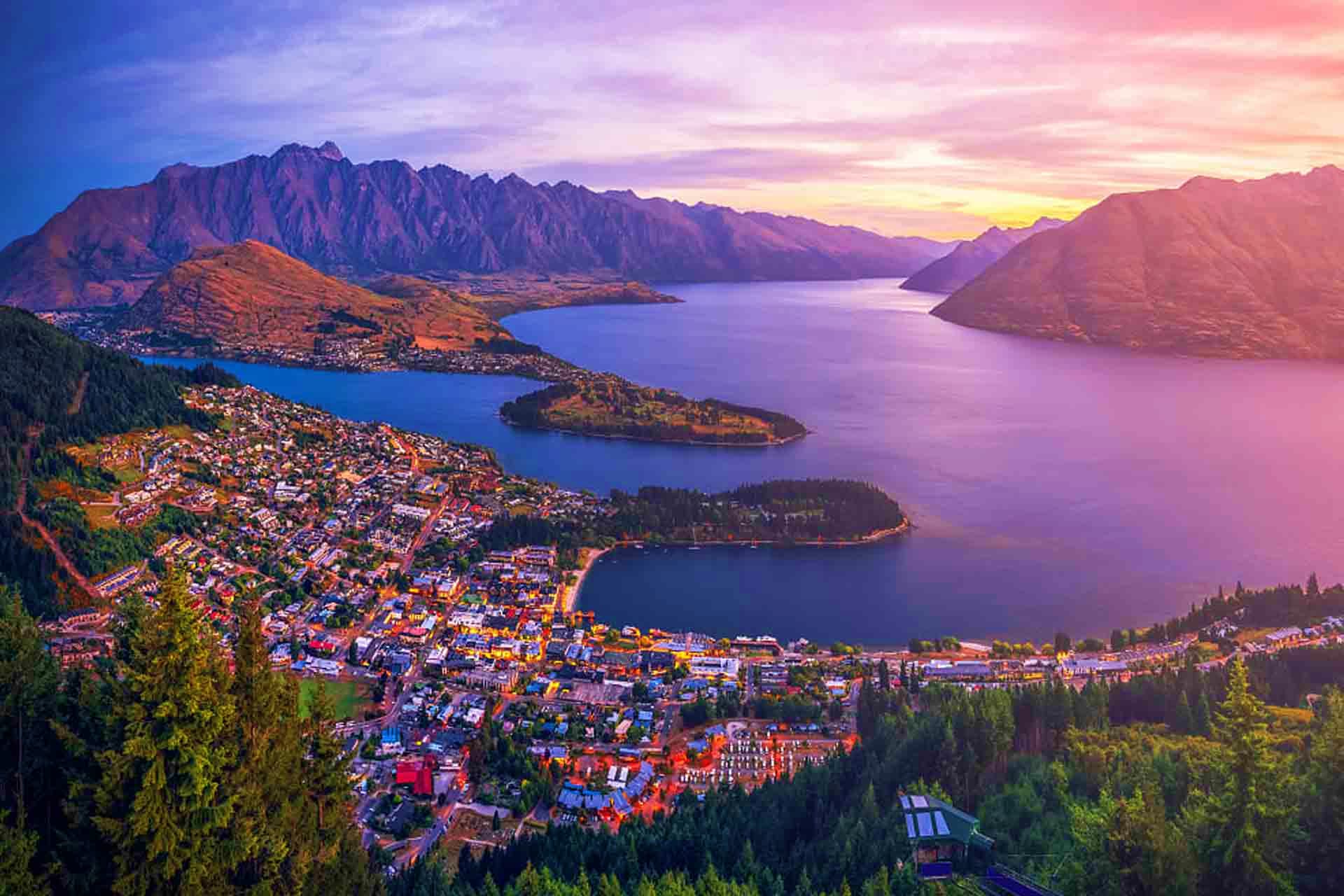 Aerial sunset view of Queenstown, New Zealand with Lake Wakatipu, surrounding mountains and forested hills at dusk